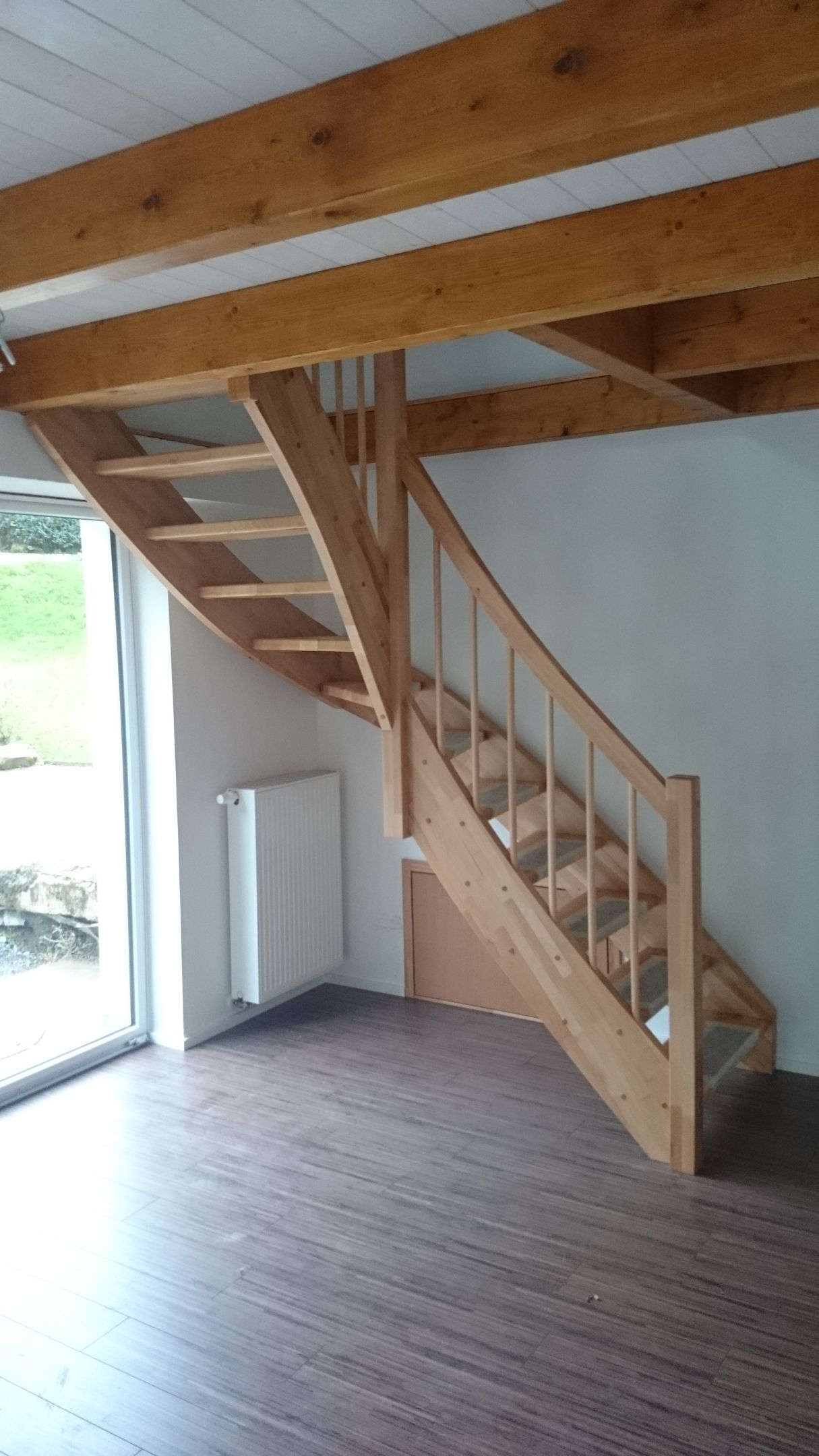 Eine hölzerne Innentreppe unter freiliegenden Balken in einem loftartigen Zimmer mit dunklem Boden und weißen Wänden
