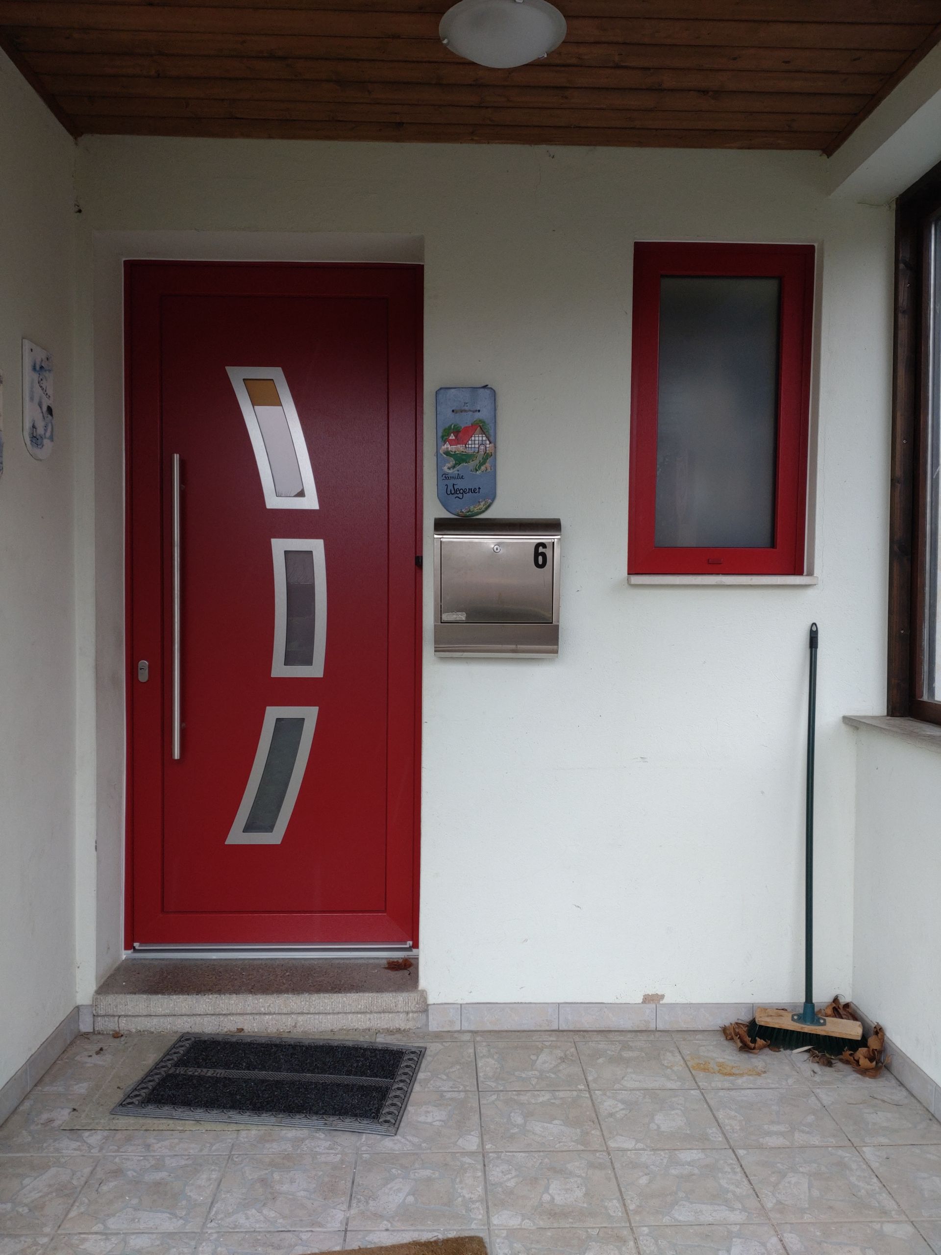 Rote Haustür mit Briefkästen und einem schmalen Fenster daneben in einem weißen Eingangsbereich