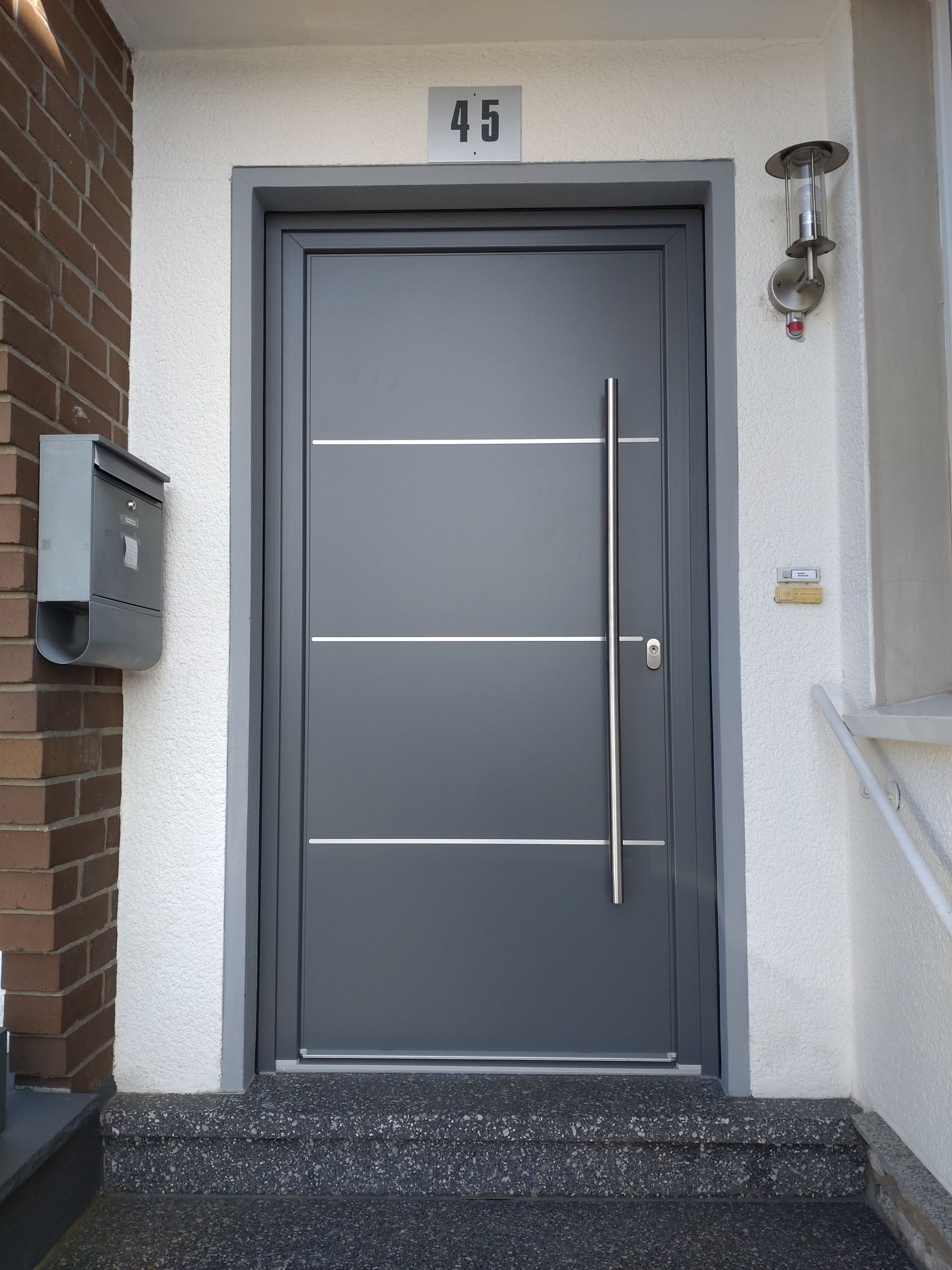 Graue Haustür mit langem, senkrechtem Griff, Hausnummer 45 und Wandbriefkasten am Eingang