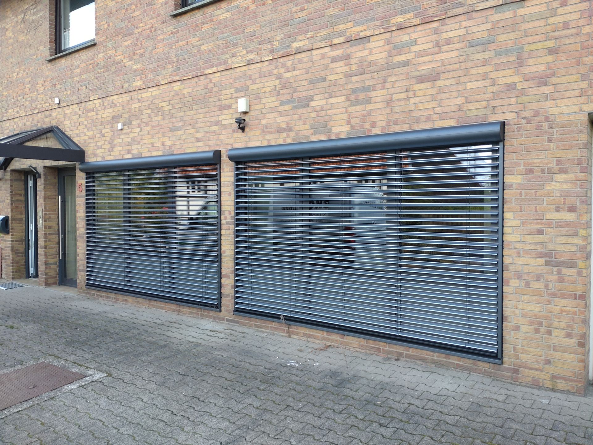 Backsteinfassade mit großem, dunkelblauem Lamellenfenster oder Garagentoren auf einem gepflasterten Gehweg