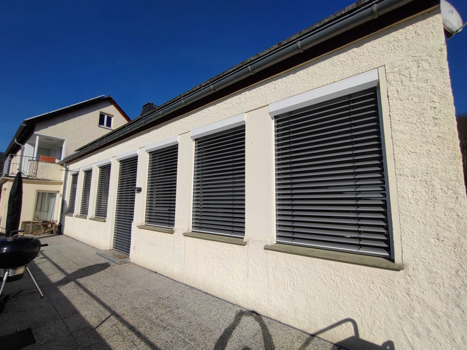 Weißes Stuckhaus mit mehreren dunklen Fensterläden an einer sonnigen Straße.