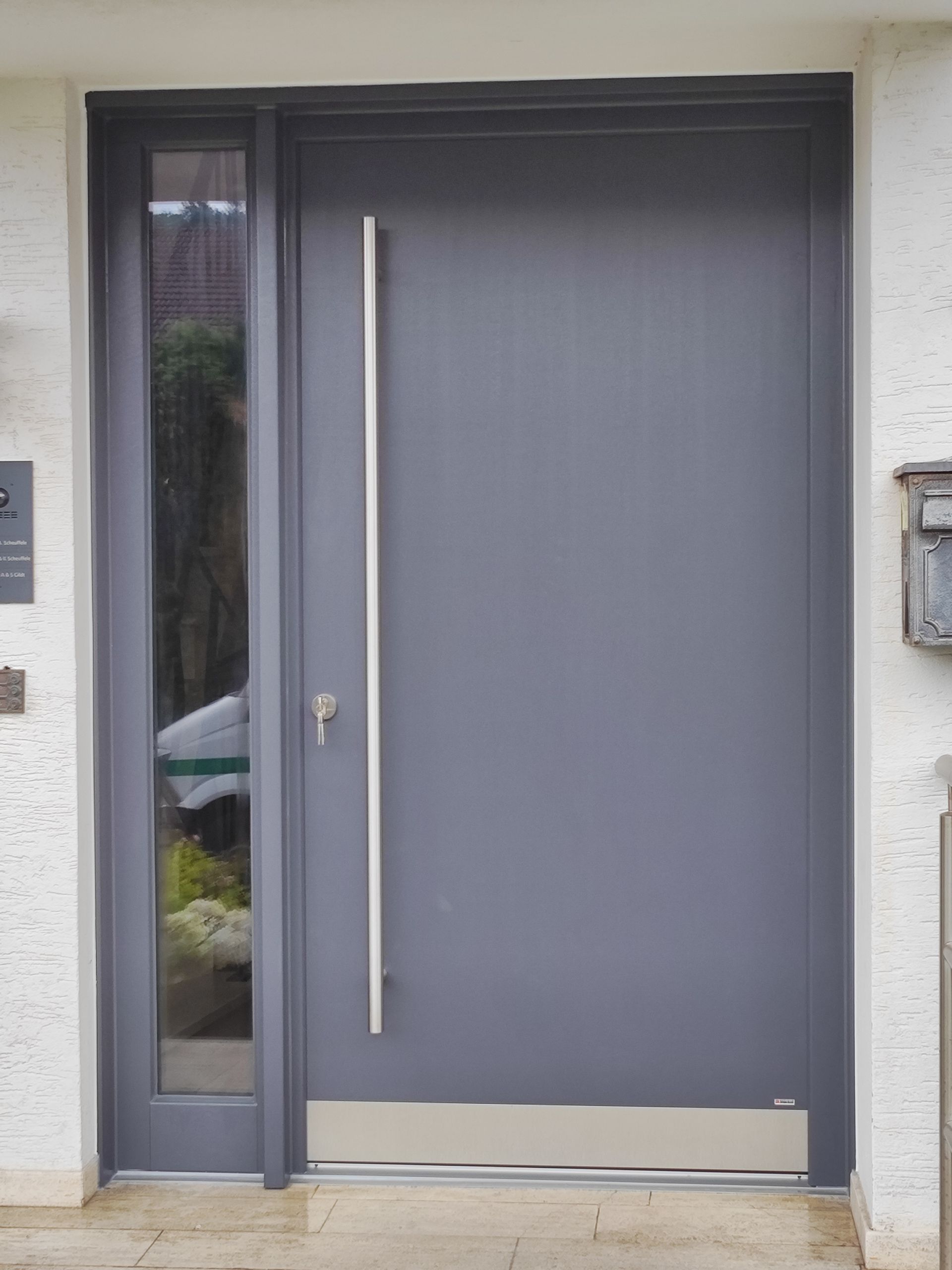 Moderne graue Haustür mit langem, vertikalem Griff und schmalem Seitenfenster in einer weißen Außenwand