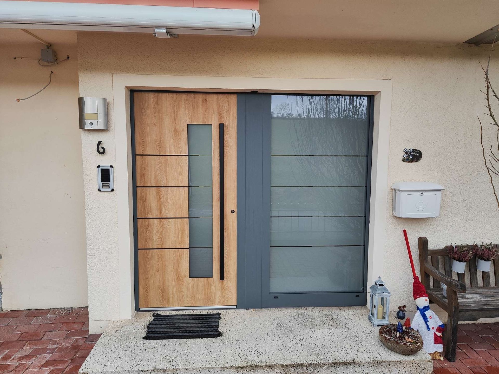 Vordereingang mit einer Tür aus Holz und Milchglas, Fußmatte und Topfpflanzen auf einer gefliesten Veranda.