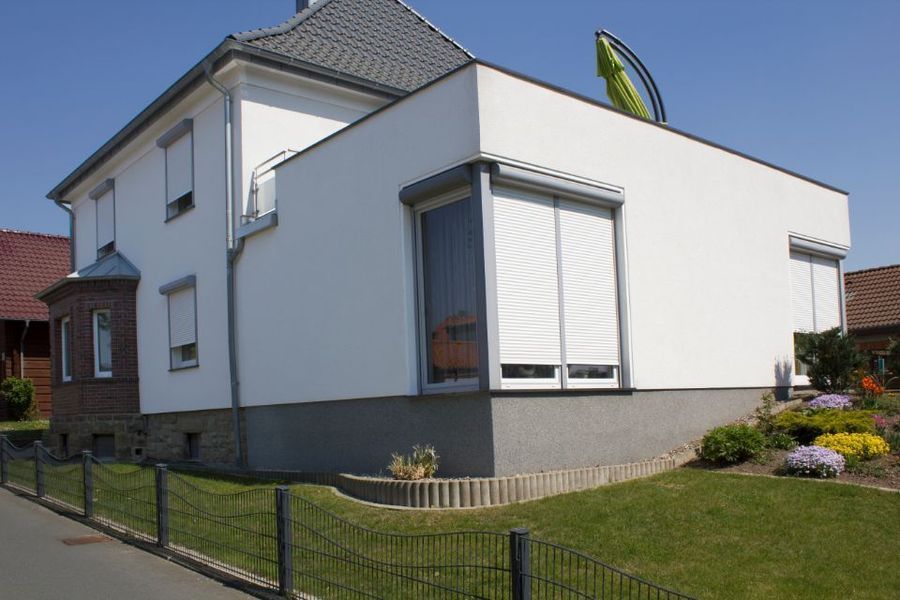 Modernes weißes Haus mit grauem Dach, umzäuntem Rasen und Blumenbeet an einem sonnigen Tag