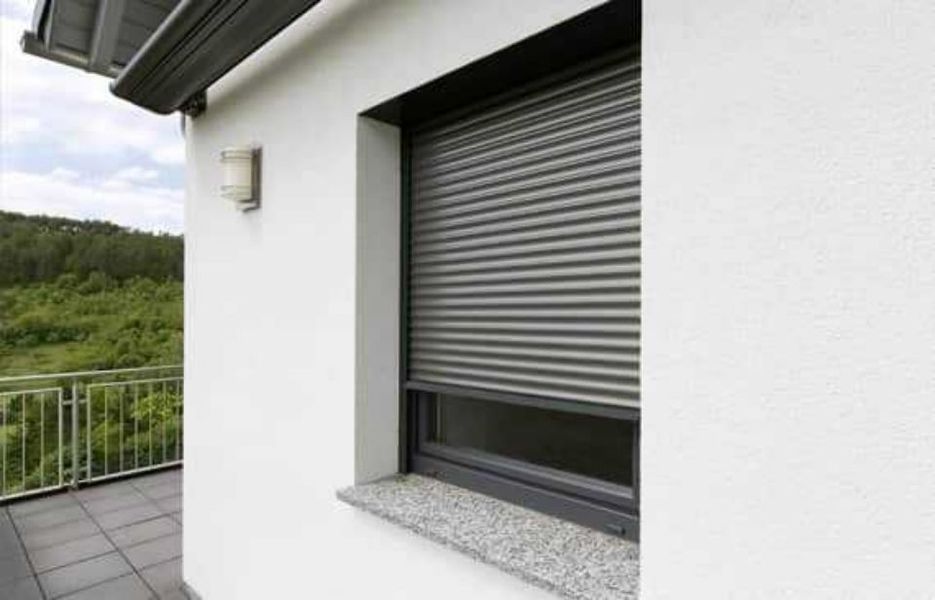 Hausfassade mit geschlossenem grauen Rollladen über einem Fenster und sichtbarem Balkongeländer