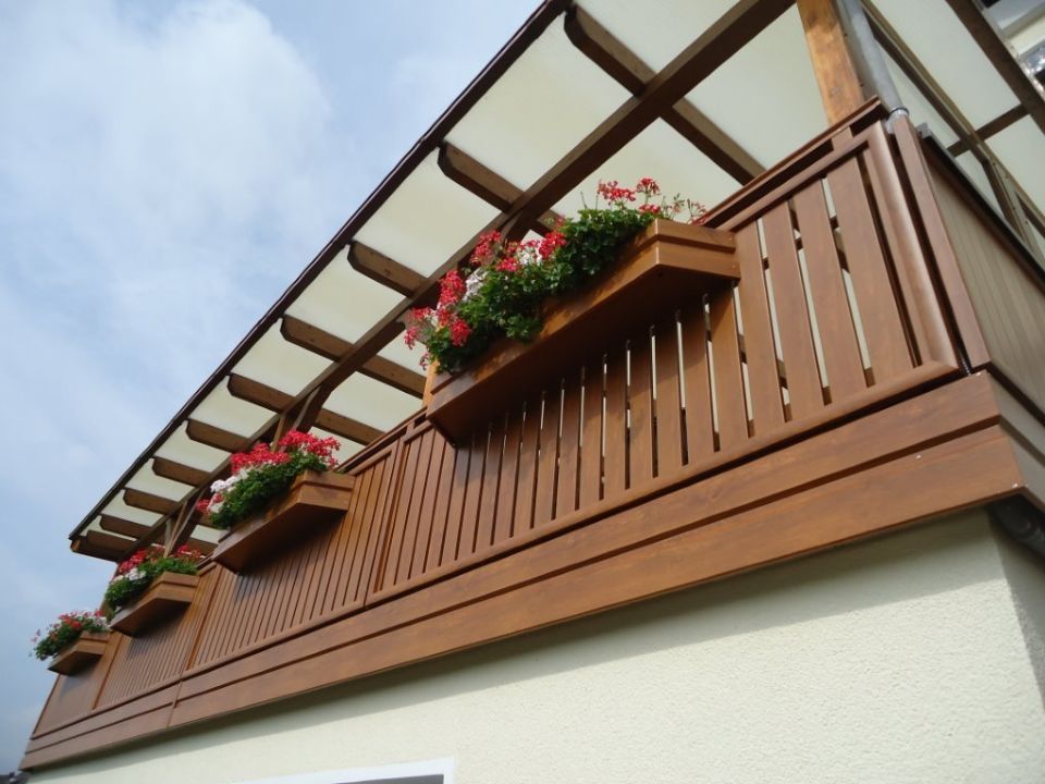 Holzbalkon mit Blumenkästen und weißer Verkleidung vor blauem Himmel