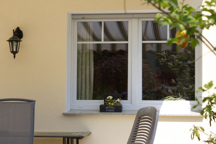 Cremefarbene Hauswand mit Fenster, Außenlampe und Terrassenstühlen auf einer sonnigen Terrasse