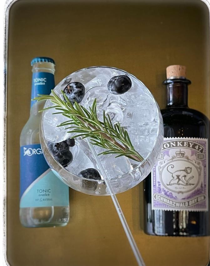 Une bouteille de gin tonic à côté d'un verre de gin aux myrtilles et au romarin.