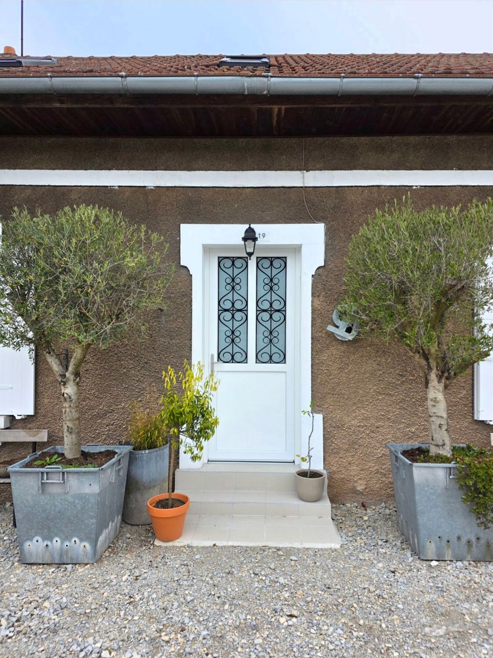 Porte d’entrée blanche avec des vitraux décoratifs, encadrée par deux arbres taillés dans des jardinières grises et une petite plante en pot sur le perron.