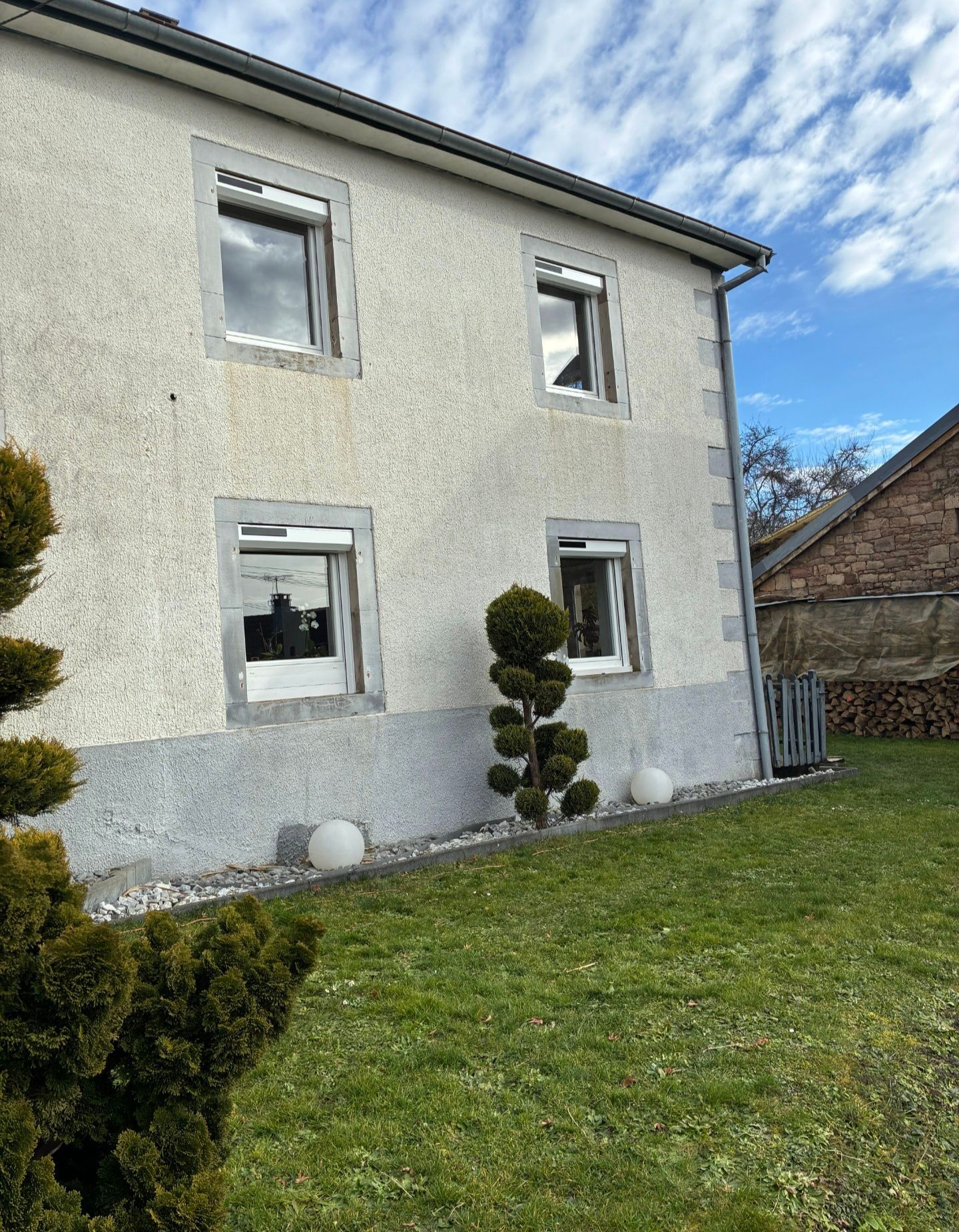 Maison crème à deux étages, aux fenêtres bordées de blanc, avec une pelouse verte et un ciel partiellement nuageux.