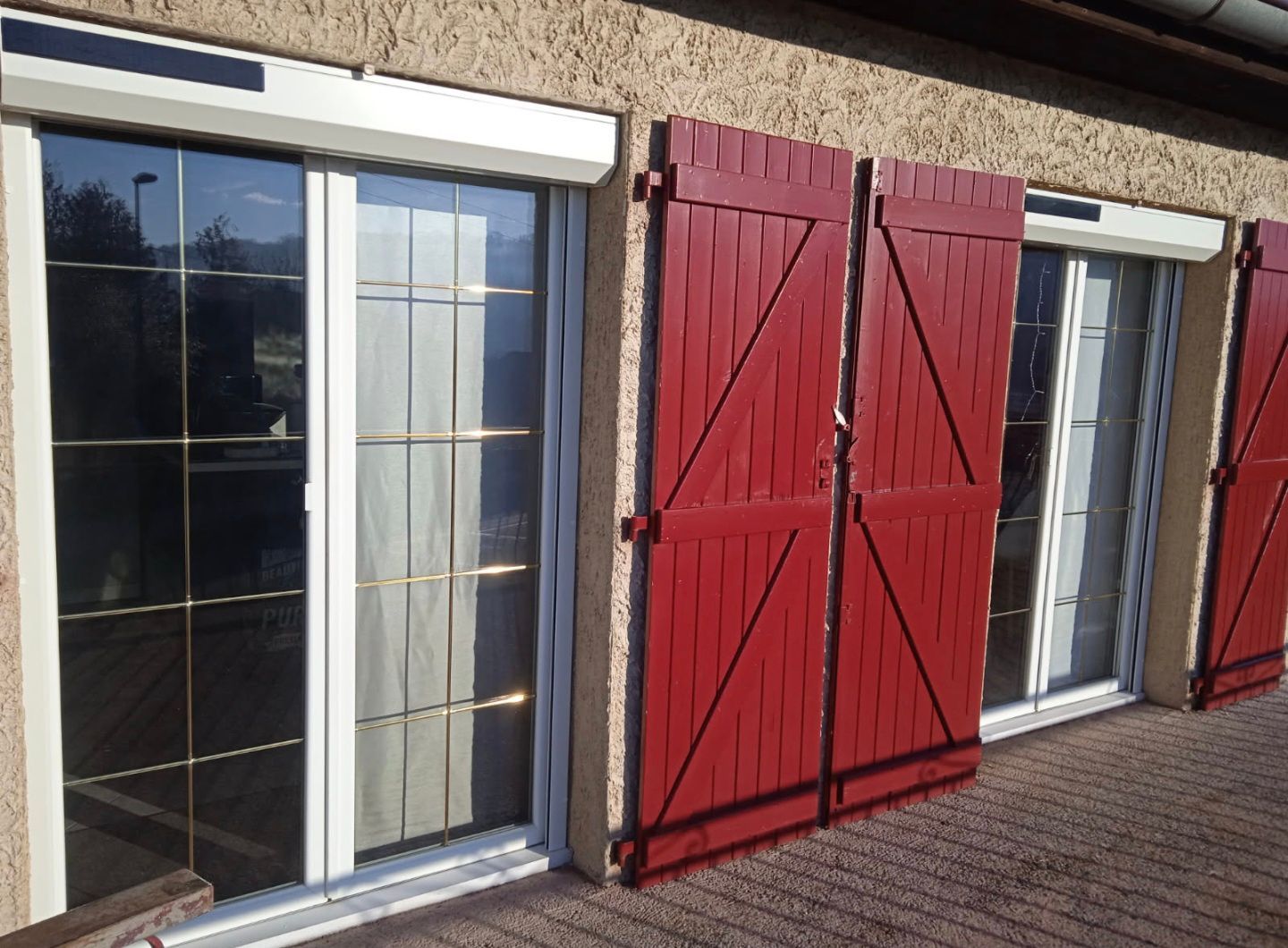 Volets rouges en bois fermés entre deux portes vitrées, sur un patio en briques