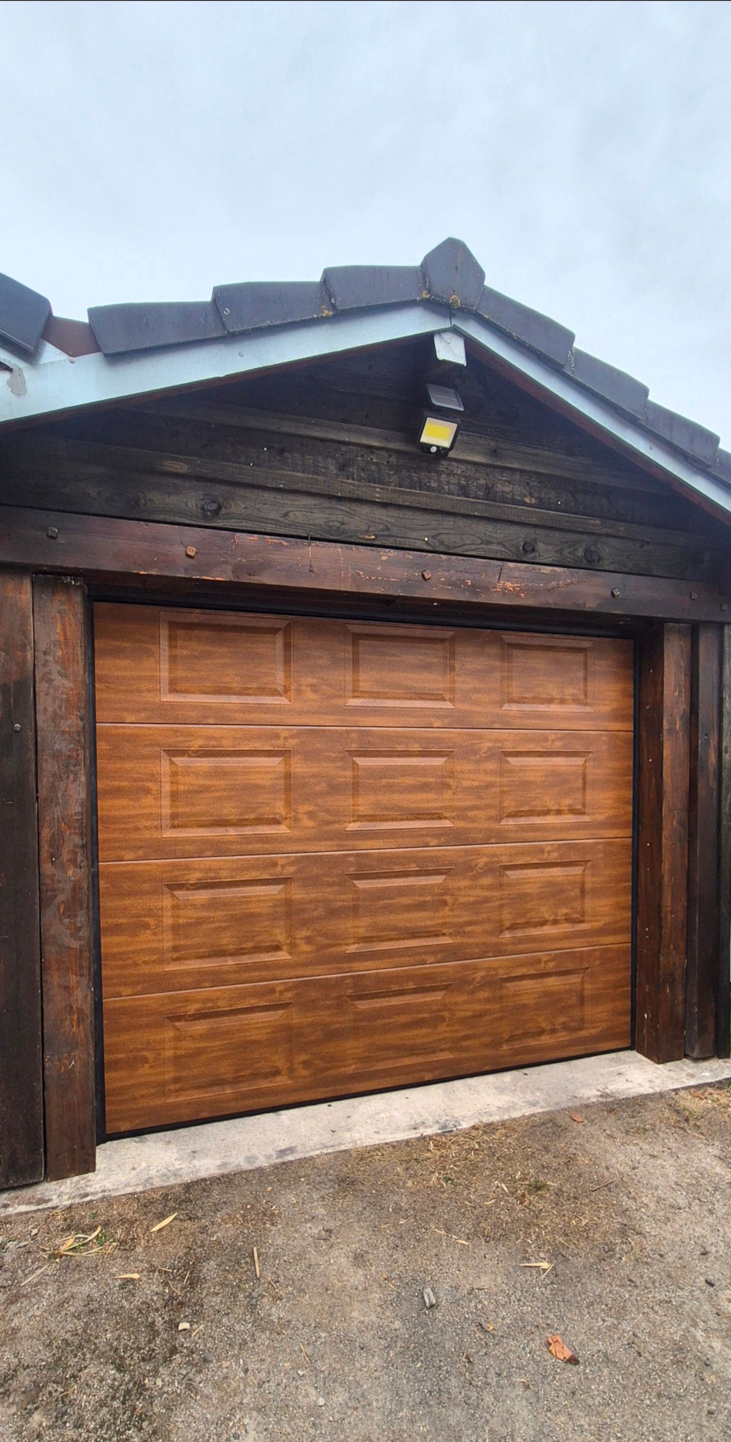 Porte de garage en bois sur un bâtiment en bois sombre, avec une allée en gravier et un ciel couvert.