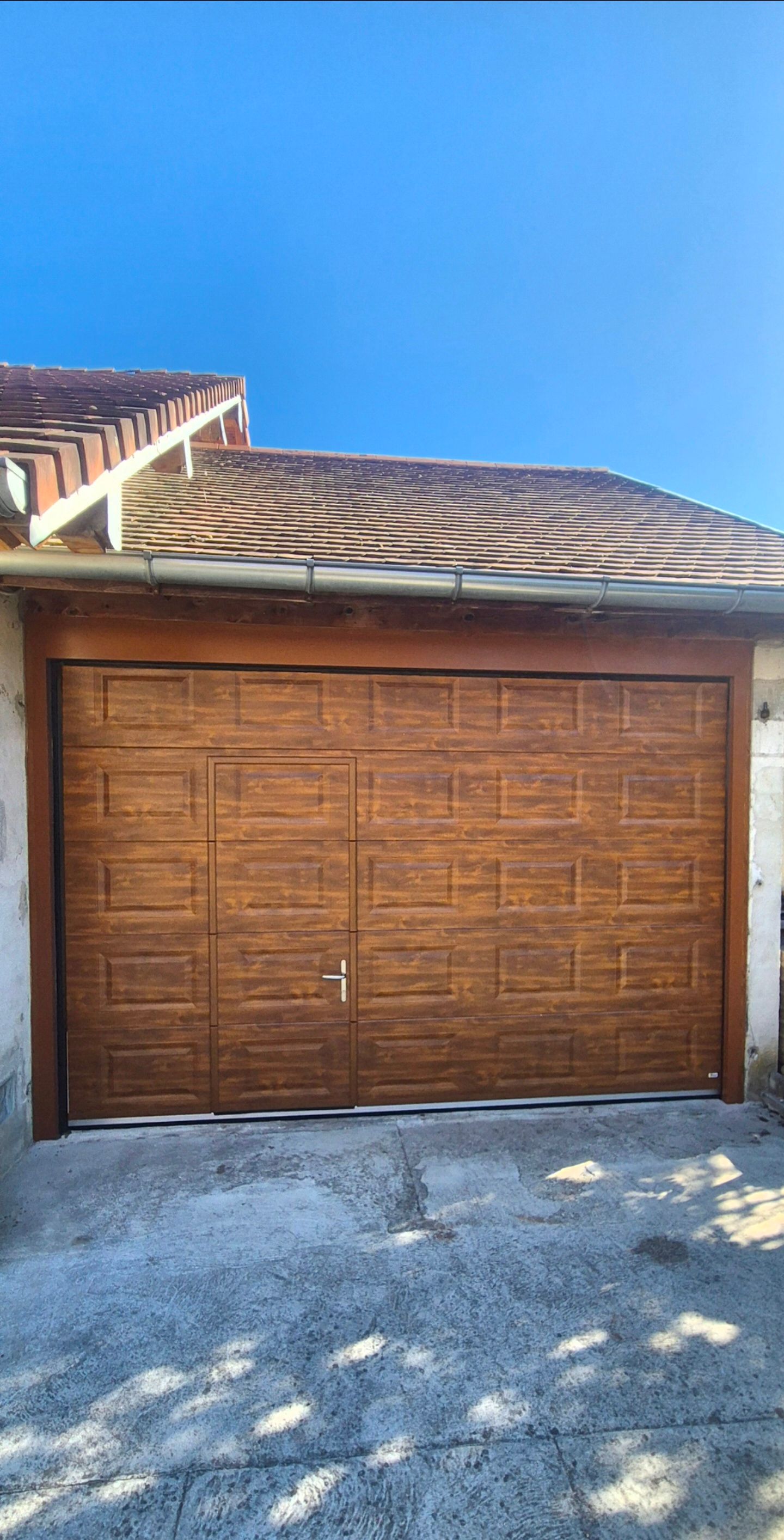 Porte de garage en bois sous un toit à motifs, avec un ciel bleu et une allée devant.