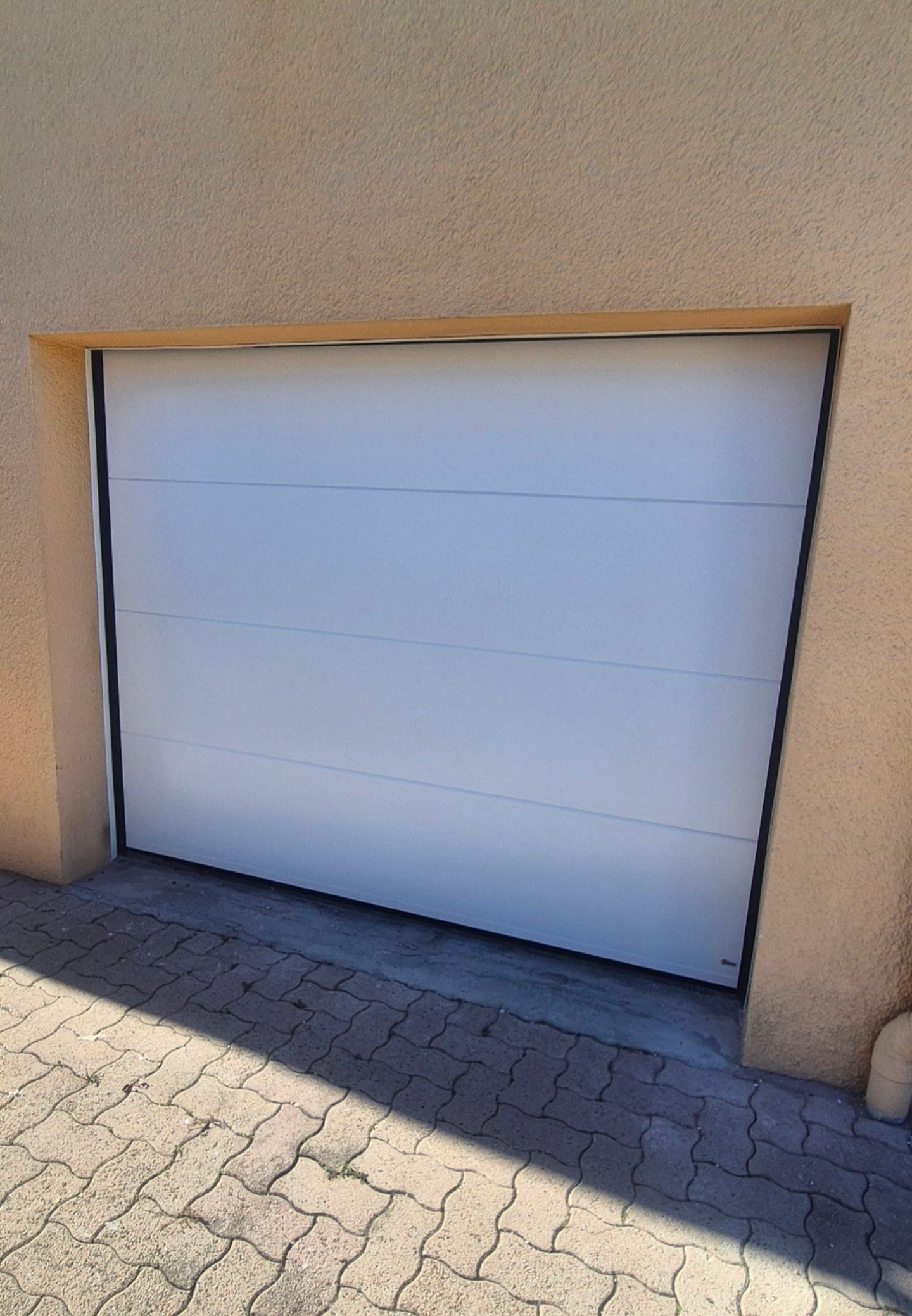 Porte de garage blanche intégrée dans un mur en stuc beige, avec une allée pavée de briques devant.