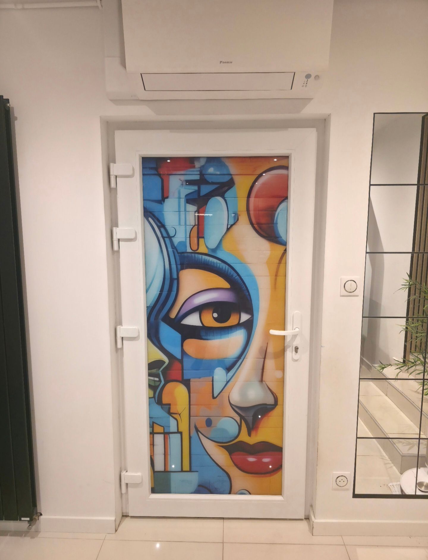 Fresque murale représentant un visage abstrait et coloré sur une porte dans un couloir, avec un miroir mural et un climatiseur au-dessus