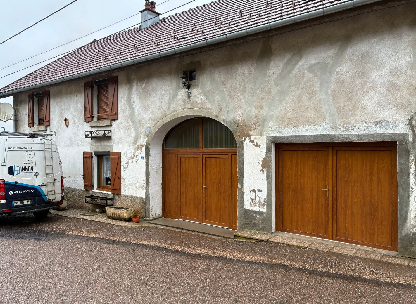 Façade d’une maison rurale en pierre avec des portes en bois, de petites fenêtres et un van stationné dans une rue gravillonnée