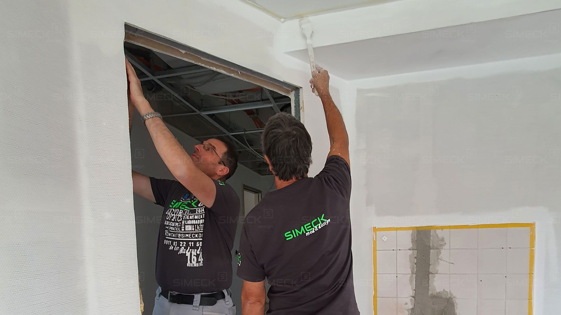 Zwei Männer arbeiten in einem Zimmer an einem Türrahmen.