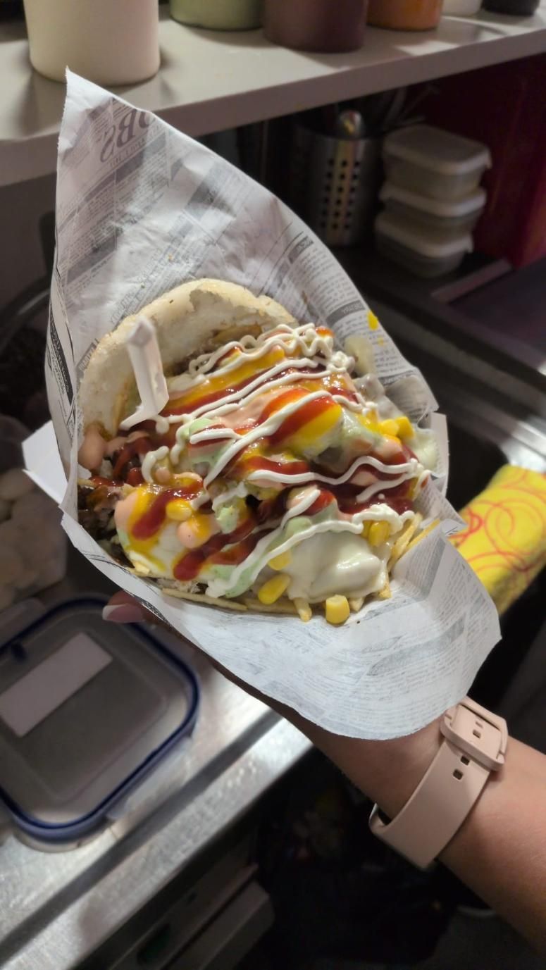 Una mano sostiene un taco relleno con salsas rociadas en una cocina o área de preparación de alimentos.
