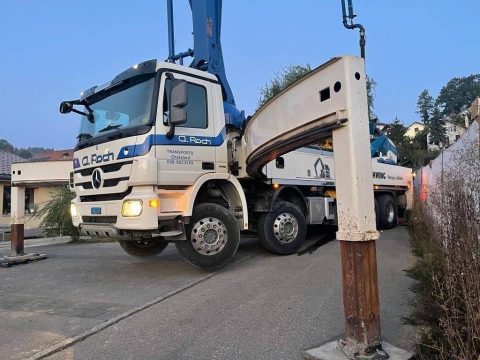 Camion-grue bleu effectuant une manœuvre à l'entrée d'un chantier, à côté d'une barrière blanche et d'une haie.