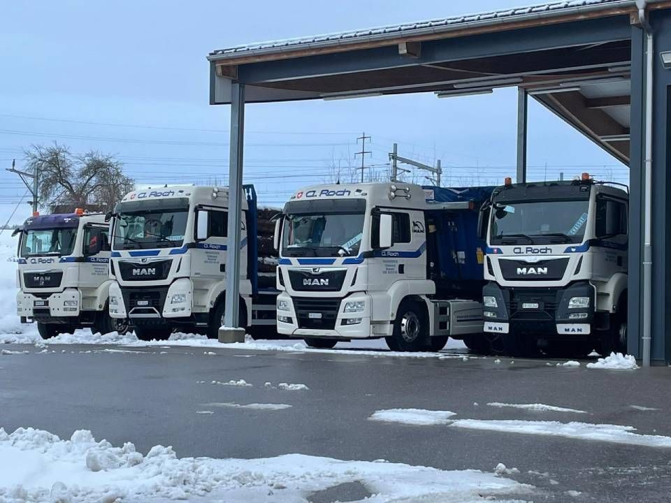 Cinq camions MAN blancs stationnés sous un abri métallique sur un terrain enneigé