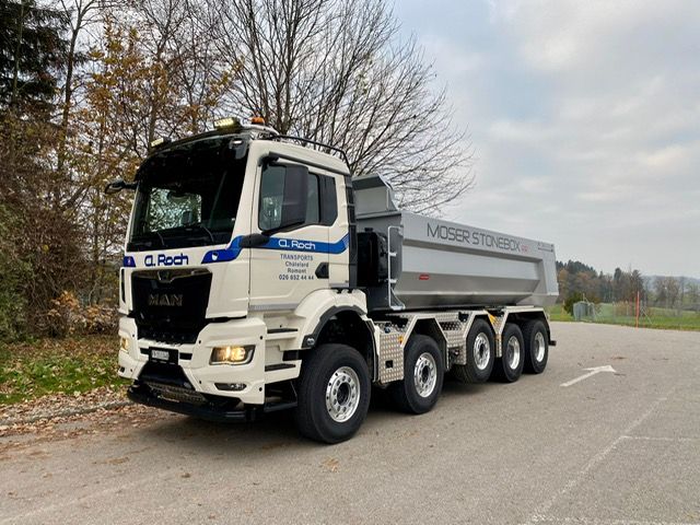 Un camion grumier blanc chargé de grumes coupées roule sur un chemin de terre sous un ciel bleu clair.