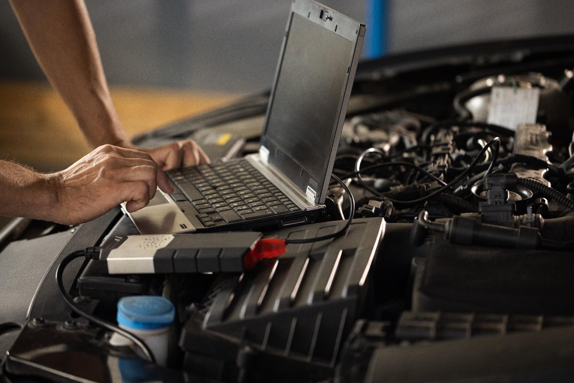Ein Mechaniker tippt in einer Garage neben einem offenen Motorraum auf einem Laptop.