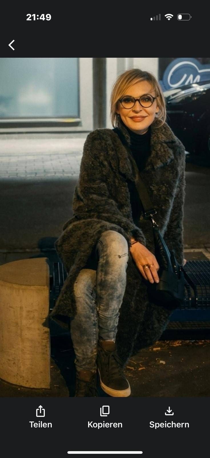 Eine Person mit Brille, die einen dunklen Kunstpelzmantel, einen schwarzen Rollkragenpullover und helle Jeans trägt, sitzt nachts im Freien.