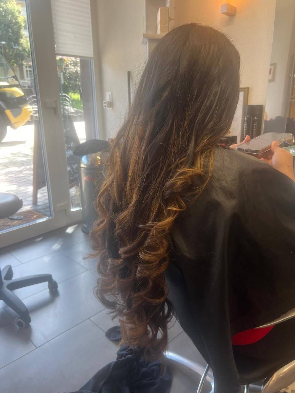 Eine Person sitzt in einem Friseurstuhl. Sie hat langes, dunkles, gewelltes Haar mit goldbraunen Strähnen.