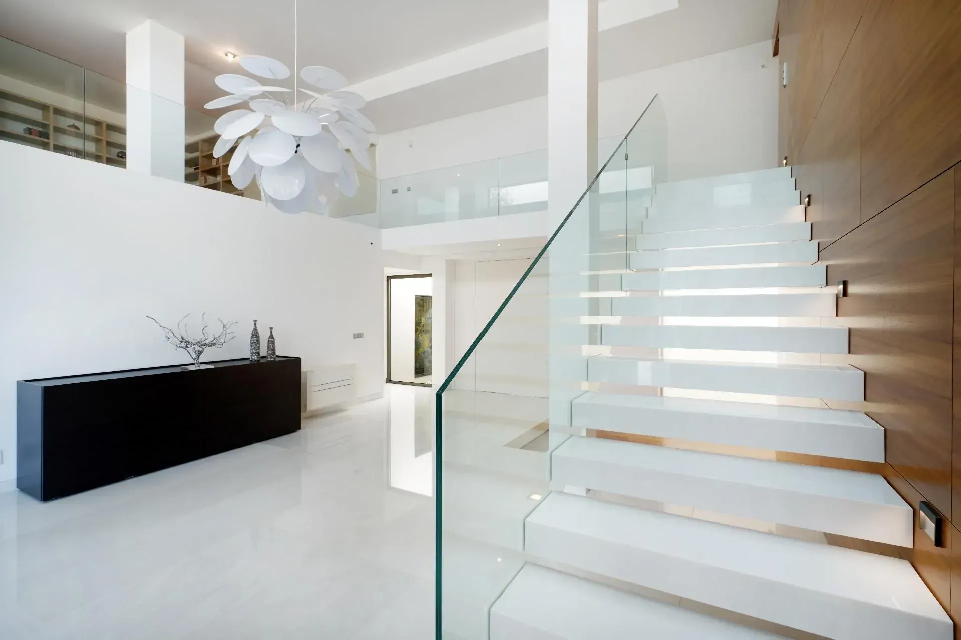 Moderne weiße Treppe mit Glasgeländer. Weiße Wände, Holzvertäfelung, schwarzer Schrank und abstrakter Kronleuchter.