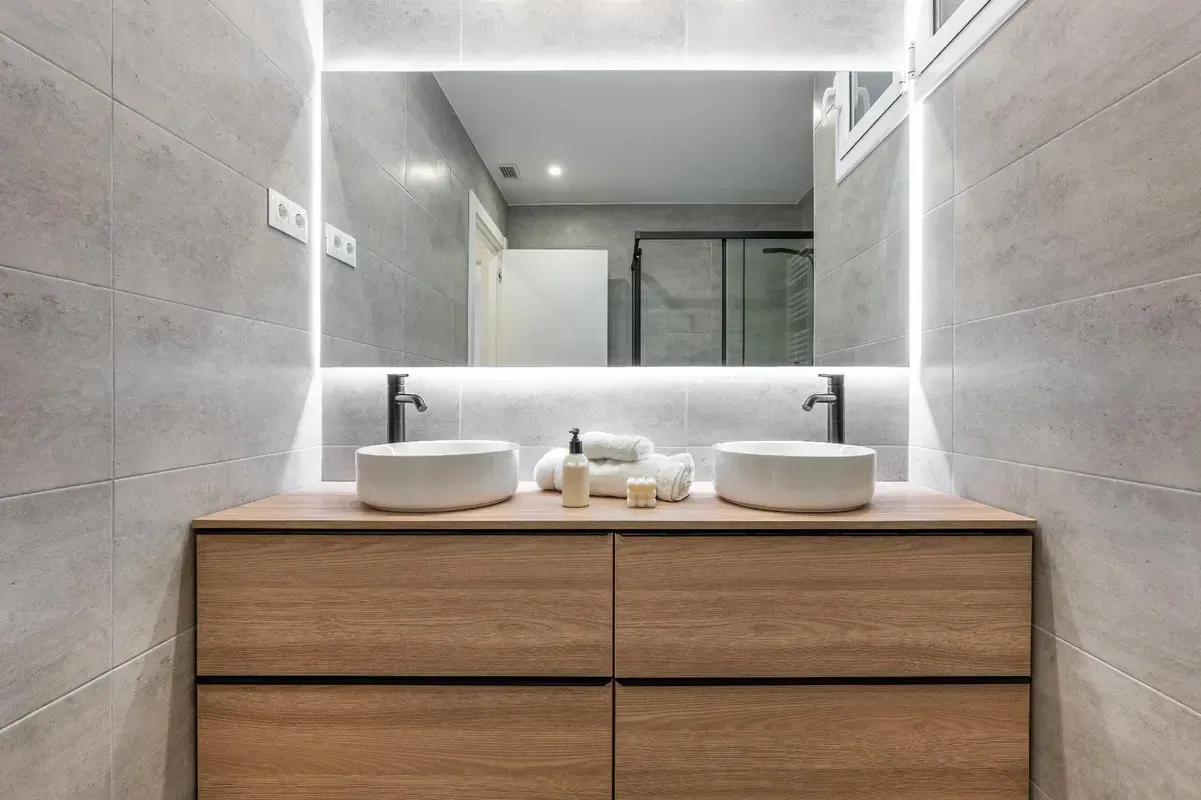 Modernes Badezimmer mit Waschtisch aus Holz, zwei weißen Waschbecken und einem beleuchteten Spiegel.