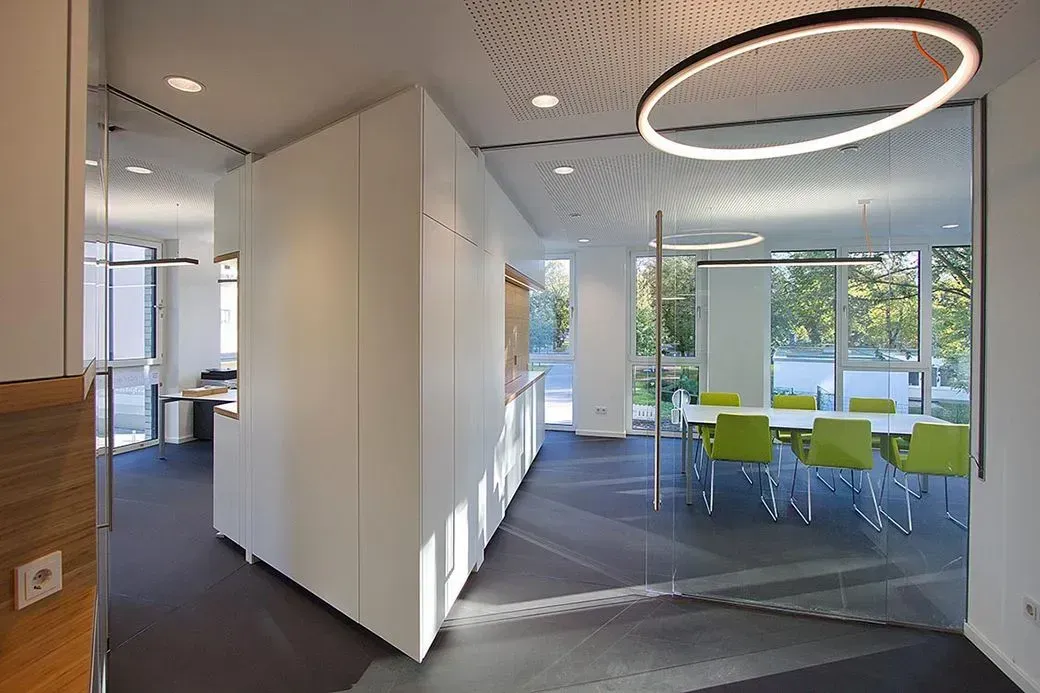 Modernes Bürointerieur mit Glaswänden, weißen Schränken und einem Konferenzraum mit grünen Stühlen.