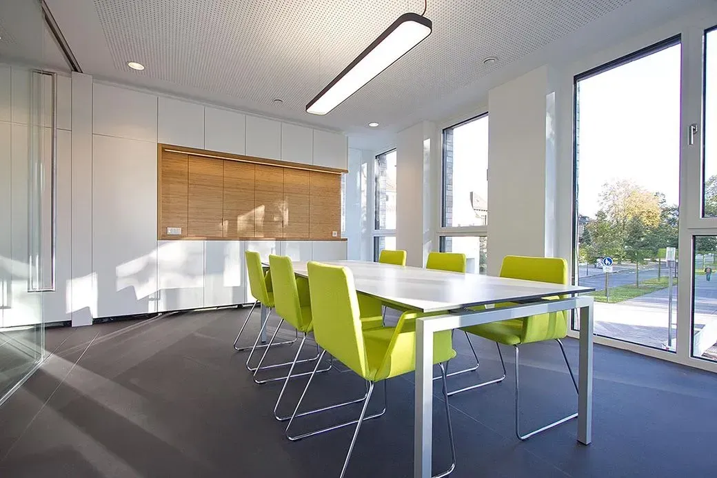 Besprechungsraum mit weißem Tisch, grünen Stühlen, großen Fenstern und einer holzgetäfelten Wand.