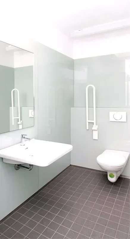 Barrierefreies Badezimmer: weißes Waschbecken und WC, grauer Fliesenboden, hellblaue Wände, Haltegriff, großer Spiegel.