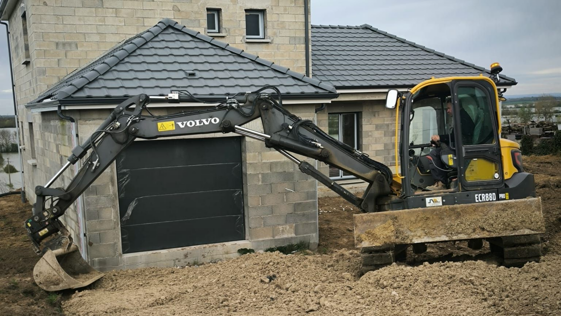 Une pelleteuse Volvo jaune creuse la terre près d'un bâtiment au toit gris avec un garage.