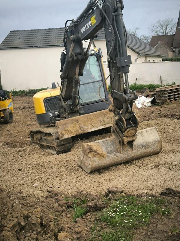 Une pelleteuse jaune et noire creuse un terrain vague, sur un chantier de construction.