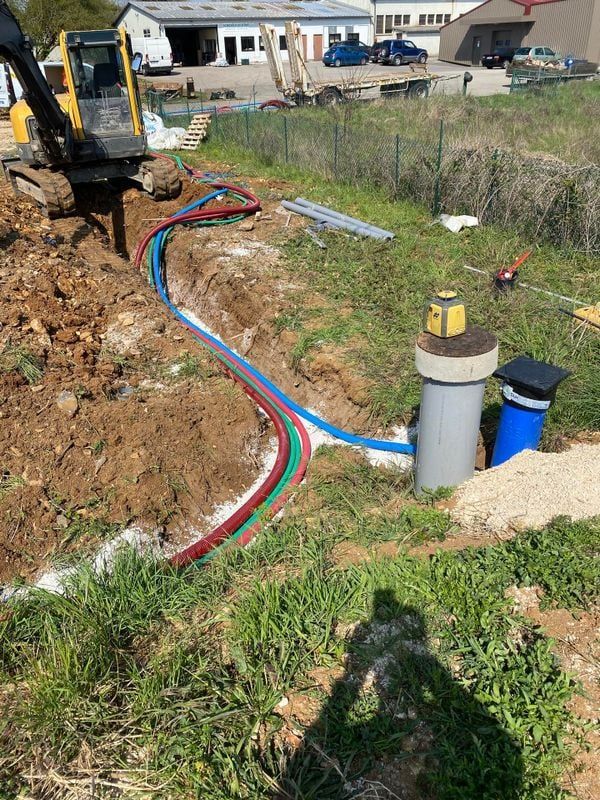 Chantier de construction avec une excavatrice et des tuyaux colorés dans une tranchée, près d'un puits et de bâtiments.