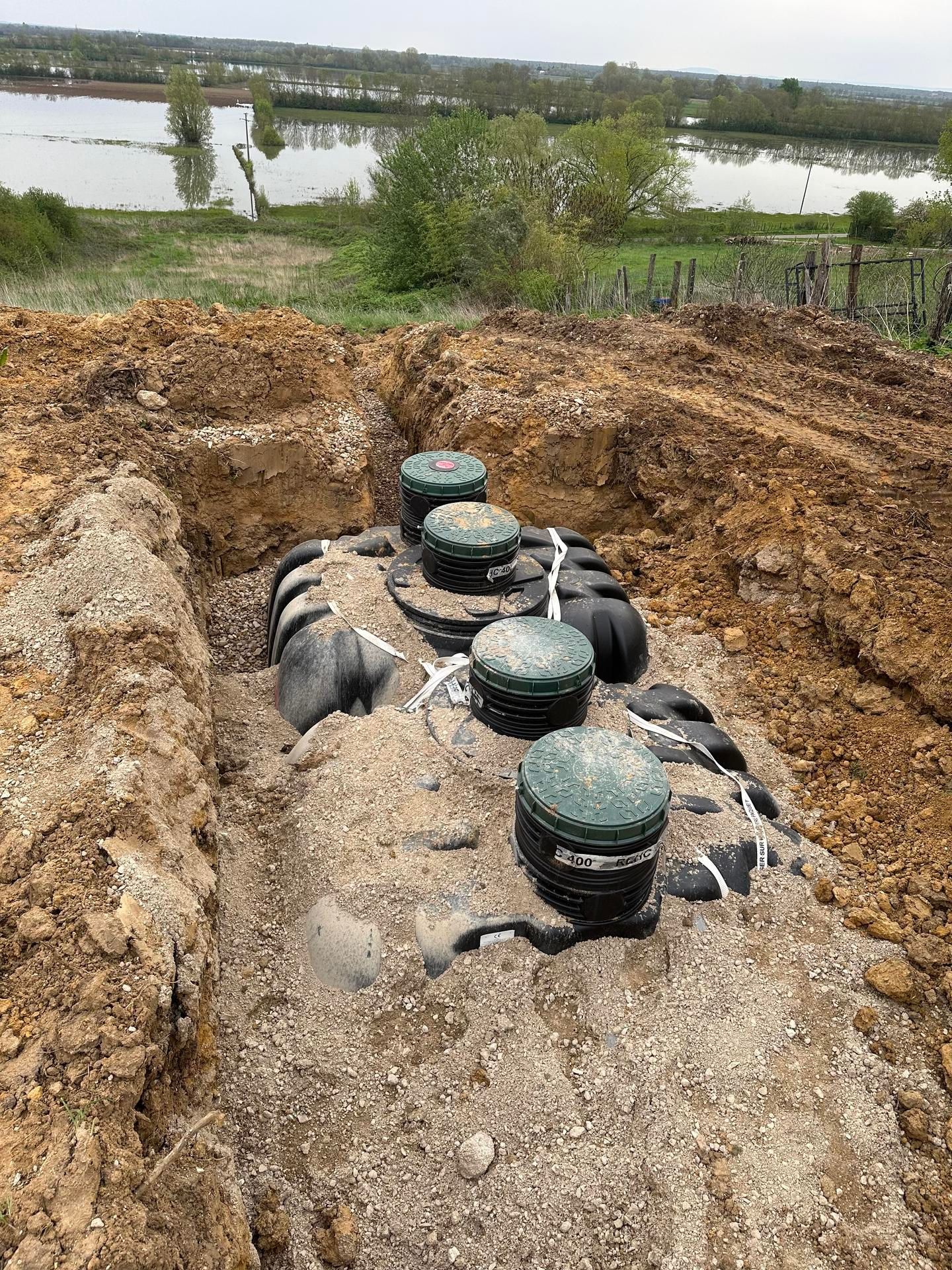 Installation d'une fosse septique dans une tranchée, avec des points d'accès à capuchon vert, en milieu rural avec un point d'eau en arrière-plan.