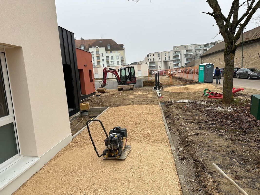 Chantier avec petite pelle mécanique, compacteur et bâtiments. Copeaux de bois sur un chemin.