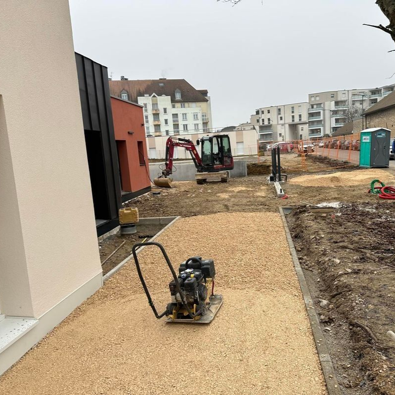 Chantier avec allée de gravier, excavatrice et compacteur. Bâtiments en arrière-plan.