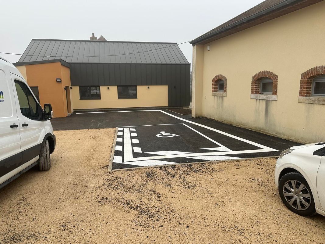 Extérieur du bâtiment avec parking goudronné ; une place accessible délimitée par des lignes blanches. Sol en gravier beige.