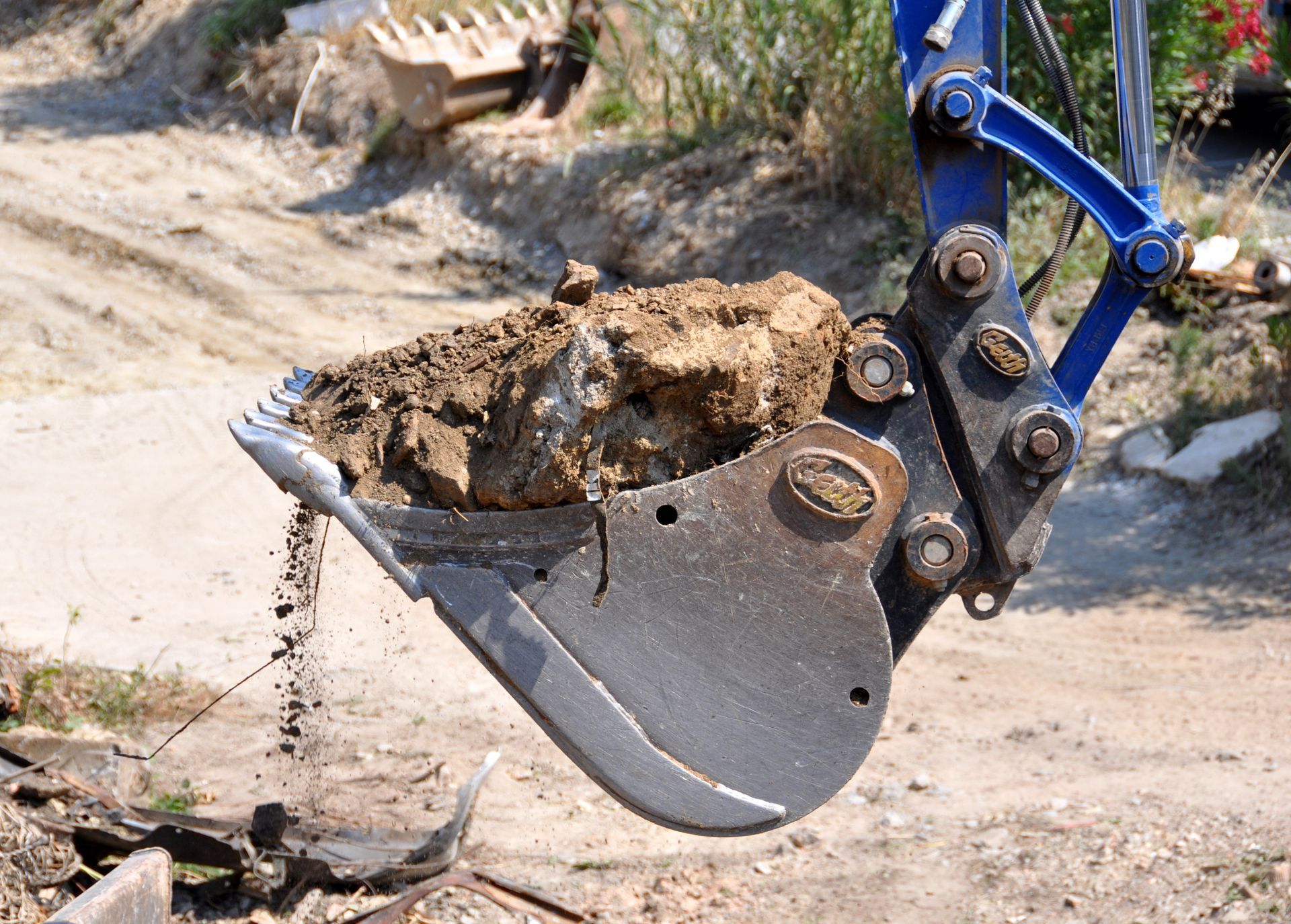 Gros plan sur le godet bleu d'une excavatrice rempli de terre et de pierres, en extérieur.