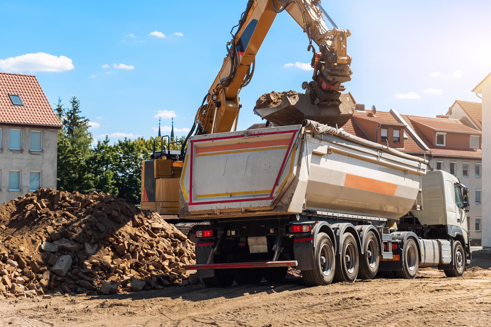 Une excavatrice charge de la terre dans un camion-benne sur un chantier de construction ; des bâtiments sont visibles en arrière-plan.