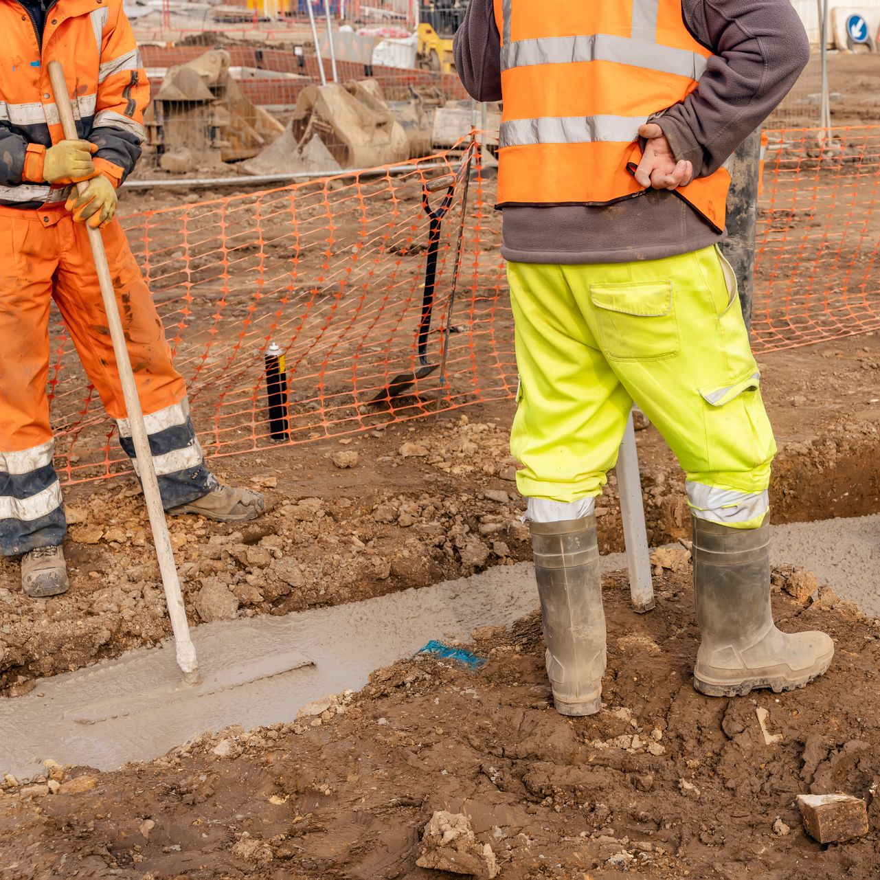 Deux ouvriers du bâtiment en tenue de sécurité sur un chantier, l'un d'eux lissant le béton avec un long outil.