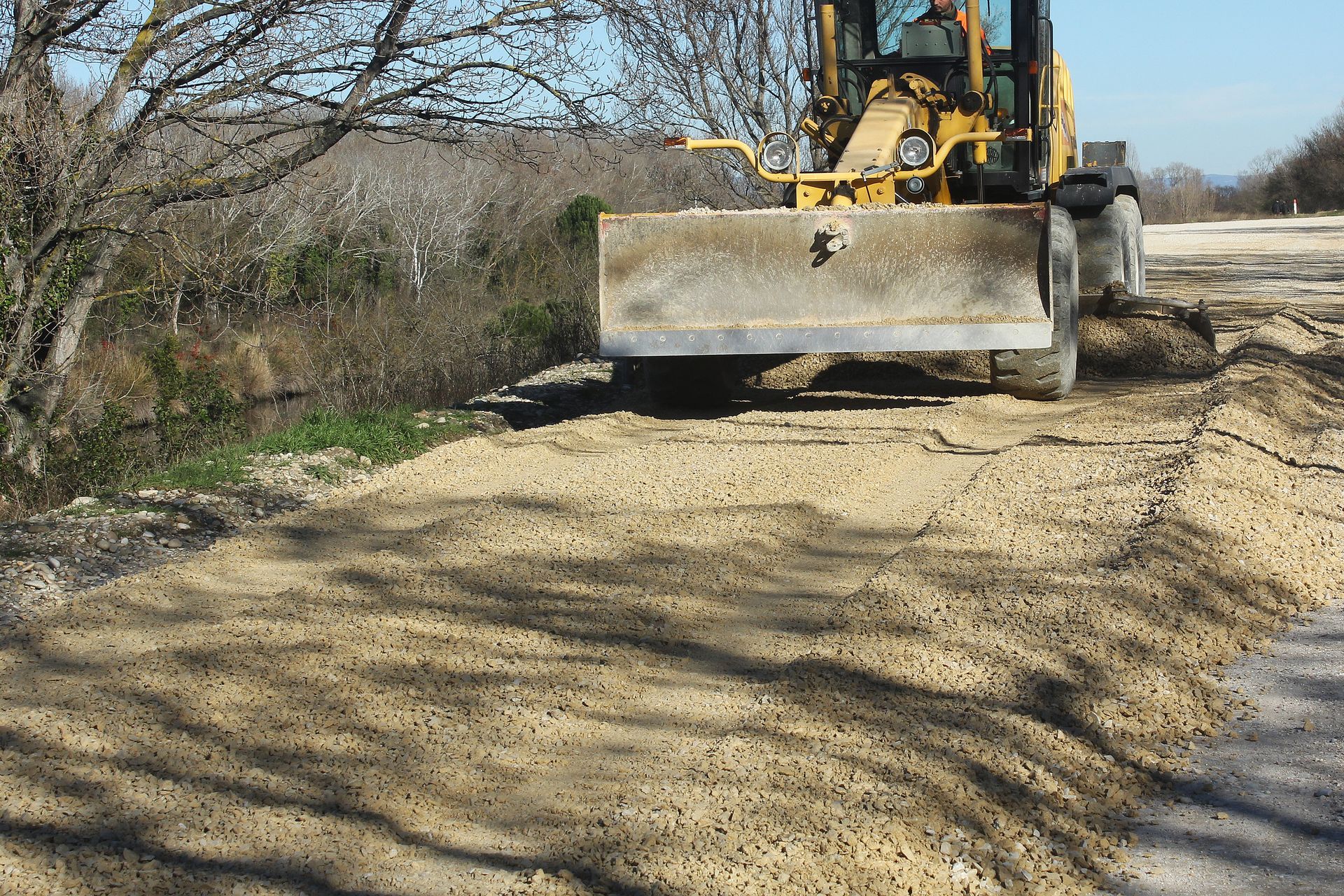Une niveleuse jaune lisse le gravier en bord de route, avec des arbres en arrière-plan.