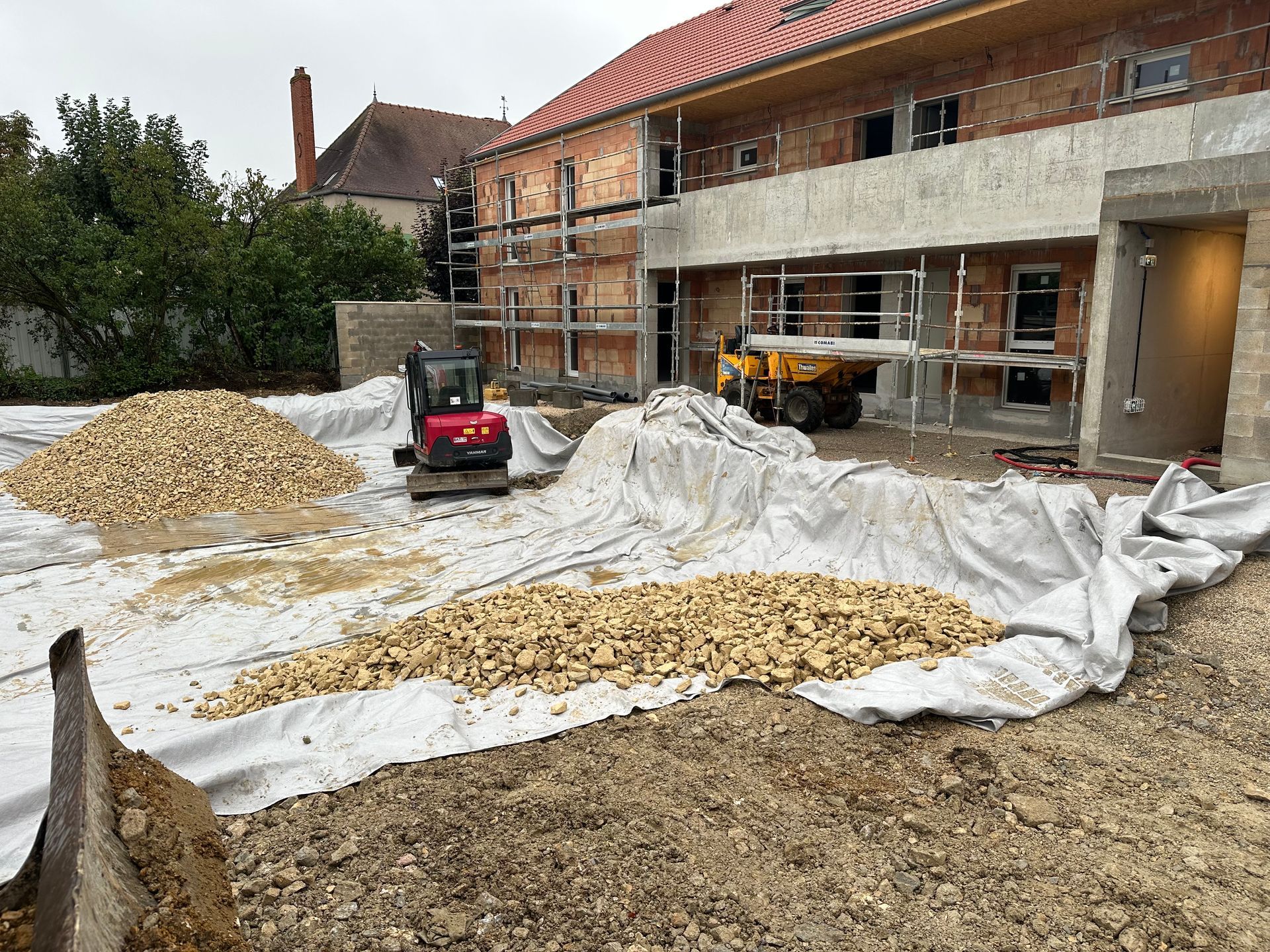 Chantier de construction avec pelle mécanique et tas de gravier, bâtiments en arrière-plan.