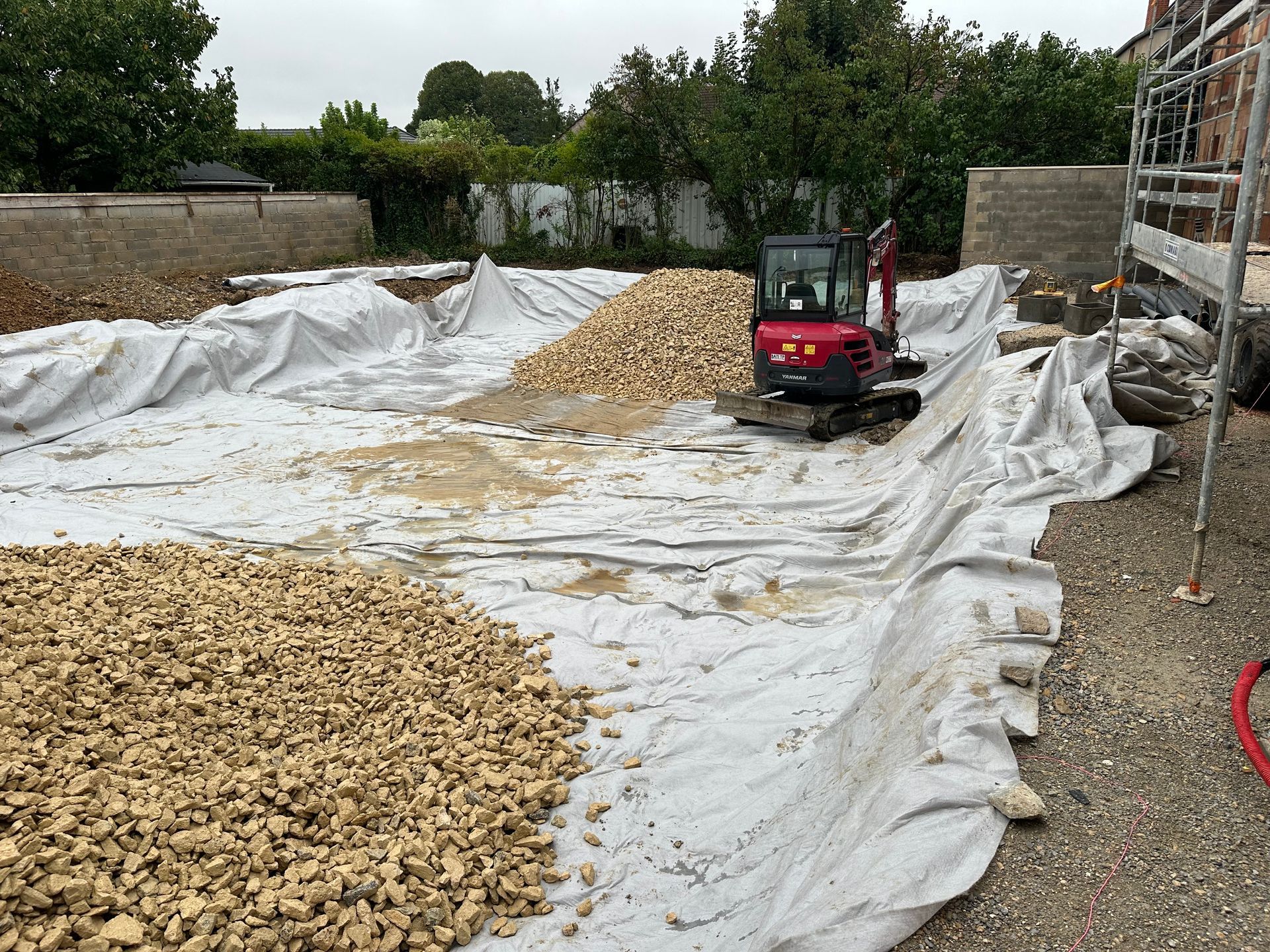 Mini-pelle sur un chantier, entourée de gravier et d'une bâche de protection.