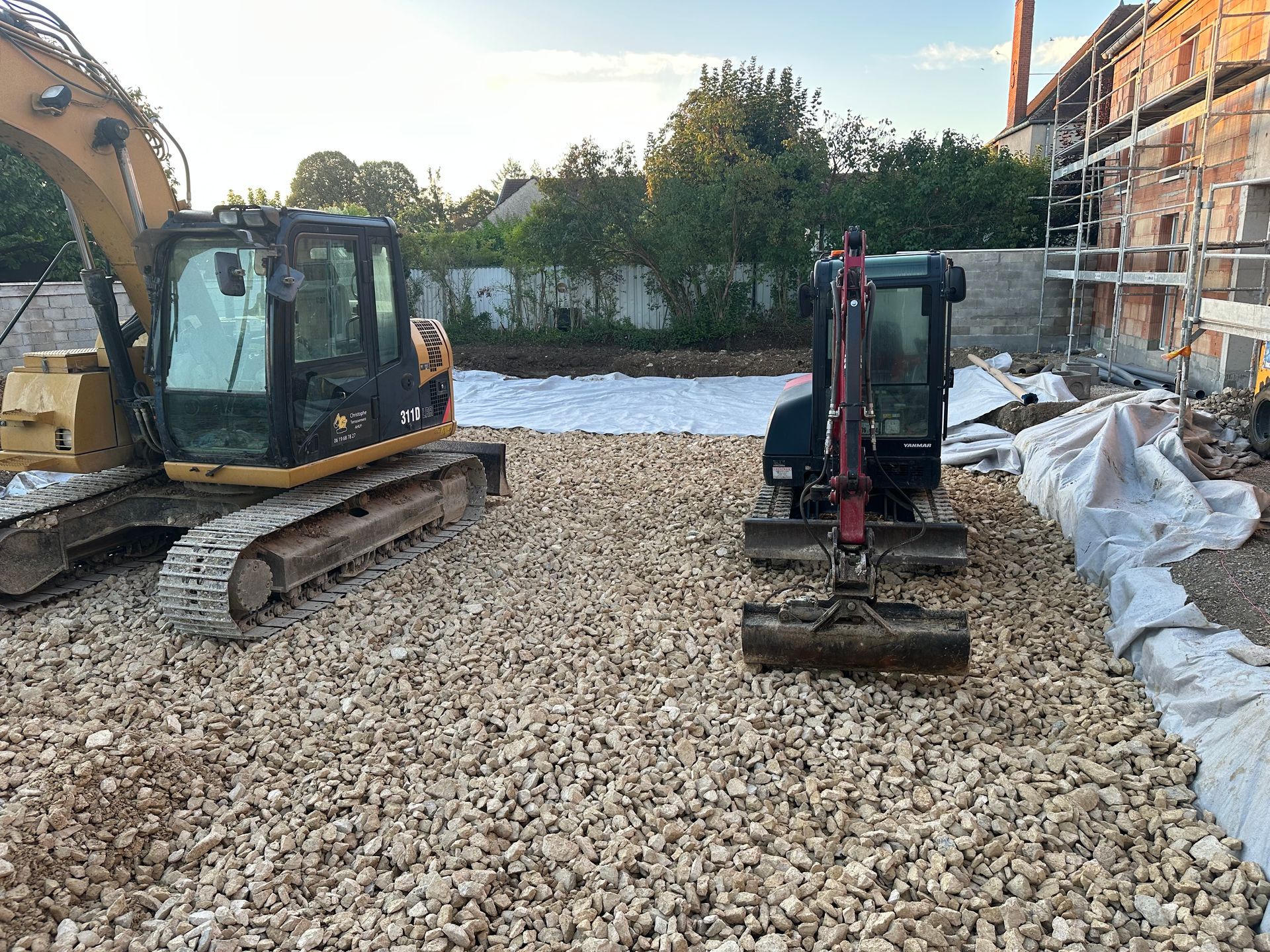 Deux excavatrices sur un lit de gravier sur un chantier de construction près d'un bâtiment en rénovation.