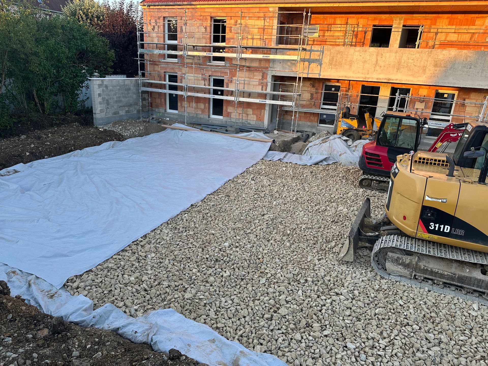 Chantier de construction avec gravier, bâche blanche et engins de chantier ; bâtiment en arrière-plan.