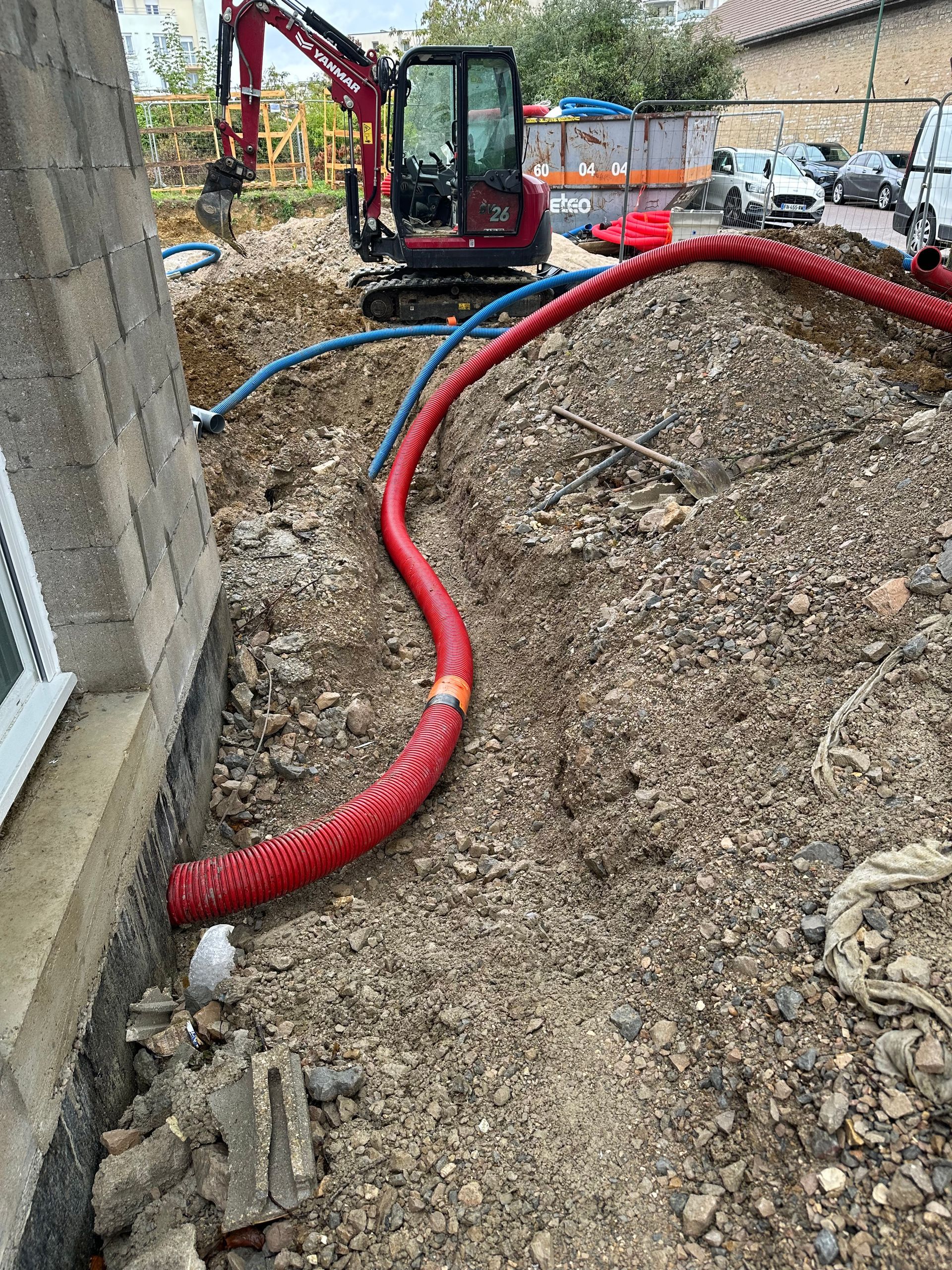 Chantier : Un conduit rouge serpente à travers un tas de terre, avec une mini-pelle en arrière-plan.