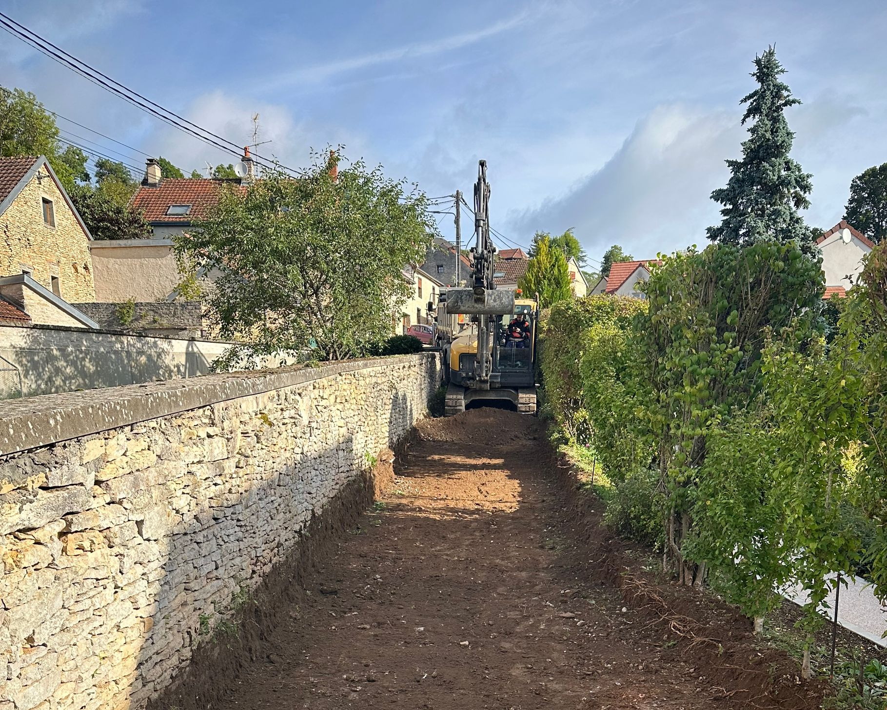 Une pelleteuse travaille sur un chemin de terre en pente, longeant un mur de pierre.
